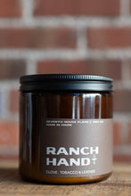 Load image into Gallery viewer, A dark amber glass jar candle with a black lid and a brown label reading 'RANCH HAND' and 'CLOVE, TOBACCO &amp; LEATHER' by Seventh House Flame &amp; Wax Co., set on a wooden surface with a blurred brick wall background.