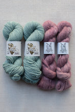 Load image into Gallery viewer, Four skeins of yarn arranged side by side on a light beige textured fabric background. The left two skeins are a muted sage green, and the right two are a dusty mauve pink.