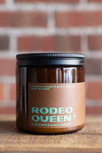 Load image into Gallery viewer, A dark amber glass candle jar with a black metal lid and a brown paper label reading 'RODEO QUEEN' and 'SPIN SUGAR &amp; BLACK CURRANT', set on a wooden surface with a blurred brick wall background.