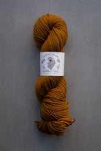 Load image into Gallery viewer, Mustard yellow skein of yarn with a label on a gray background