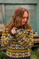 A woman wearing a brown, yellow, and white patterned knit sweater, standing outdoors in front of a rustic green wooden wall.