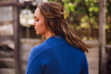 Load image into Gallery viewer, A woman with long brown hair wearing a bright blue knit sweater, shown from the back and side profile in an outdoor setting with a blurred wooden fence and greenery in the background.