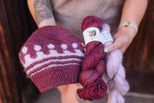 Load image into Gallery viewer, A person holds a knitted beanie in burgundy and white alongside three skeins of yarn in burgundy, pale lavender, and light pink, with a white label on the burgundy skein showing a cow illustration.