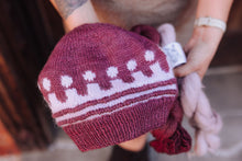 Load image into Gallery viewer, A close-up of a hand holding a hand-knit beanie in burgundy with a white geometric pattern. A small white tag is attached, and a bundle of light pink yarn is held alongside it.