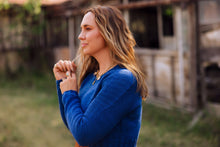 Load image into Gallery viewer, A woman with long wavy light brown hair stands outdoors in profile, wearing a textured royal blue knit sweater and a gold pendant necklace. The background shows a softly blurred rustic wooden structure and green grass.