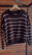 Load image into Gallery viewer, A dark brown knit sweater with horizontal light pink stripes, hanging on a wooden hanger against a red brick wall.