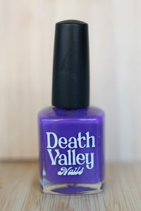 Purple nail polish bottle with black cap labeled 'Death Valley Nails' on a wooden surface.