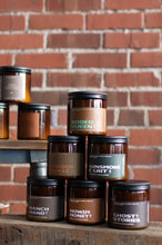 Load image into Gallery viewer, A stack of amber glass jar candles with black lids and brown labels, arranged on wooden shelves in front of a red brick wall. Visible label text includes 'Rodeo Queen', 'Cowboy Coffee', 'Gunsmoke &amp; Grit', 'Ranch Hand', 'Howdy Honey', and 'Ghost Stories'.