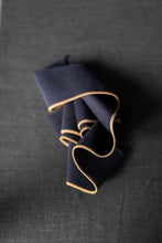 Load image into Gallery viewer, A folded navy blue fabric accessory, likely a pocket square or napkin, featuring a thin tan or gold contrast edge, displayed on a dark textured gray background.