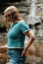 Load image into Gallery viewer, A woman with long light brown hair stands outdoors in front of a blurred waterfall, wearing a teal short-sleeve knit top and blue jeans.