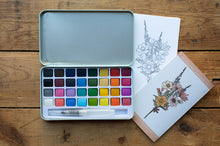 Load image into Gallery viewer, An open metal watercolor paint palette with multiple color pans and a brush, placed on a wooden surface next to two floral art cards or prints.