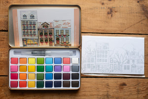 Watercolor paint set with a book of watercolor illustrations on a wooden surface
