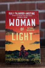 Load image into Gallery viewer, Book cover of 'Woman of Light' by Kali Fajardo-Anstine on a stand against a brick wall background.