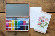 Load image into Gallery viewer, A watercolor paint palette in an open copper-toned metal tin, shown alongside a blank sheet and a floral art card on a rustic wooden surface.