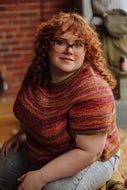 Person wearing a colorful knitted sweater sitting on a wooden floor with a brick wall background.
