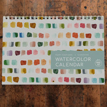 Load image into Gallery viewer, Watercolor calendar with colorful square patterns on a wooden surface
