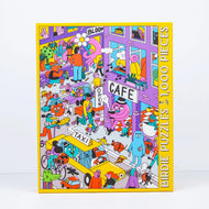 Jigsaw puzzle box with colorful illustration of a cafe scene and 'Bloom' branding.