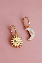 Load image into Gallery viewer, A pair of gold-toned hoop earrings, one featuring a smiling sun face charm with pearl accents and the other a crescent moon charm with a pearl, displayed on a textured pink background.