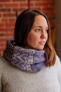 A woman wearing a chunky knit cowl scarf with a purple and gray pattern and leopard print panels, paired with a cream knit sweater against a brick wall background.