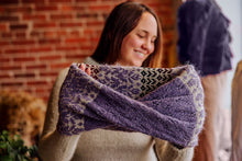 Load image into Gallery viewer, A woman smiling and holding up a chunky knit cowl in a purple and cream patterned yarn, with a brick wall background.