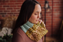 Load image into Gallery viewer, A woman wearing a chunky knit cowl neck warmer with a color-blocked design of sage green and mustard yellow with pink floral patterns, set against a warm, blurred brick wall background.