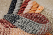 Load image into Gallery viewer, A close-up of several knitted or crocheted yarn samples in different colors arranged on a neutral textured surface. The visible colors include dark charcoal, light gray, dusty rose, peach, orange, and cream.