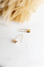 Load image into Gallery viewer, A pair of gold safety-pin style earrings with small square shield charms, displayed on a white marble surface next to dried pampas grass.