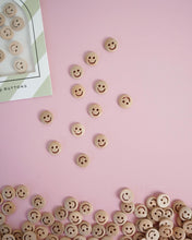 Load image into Gallery viewer, Smiley face buttons scattered on a pink surface with a box of buttons in the corner.