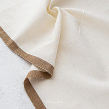 Load image into Gallery viewer, Close-up of beige fabric with brown trim on a textured surface, featuring the brand 'FAIREISM'.