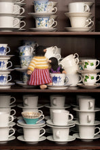 Load image into Gallery viewer, A collection of stacked white ceramic teacups and saucers on dark wooden shelves, with two small plush toy birds placed among the cups.