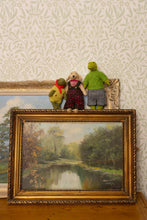 Load image into Gallery viewer, A framed landscape painting of a river scene is displayed on a wooden surface against patterned wallpaper, with three small plush stuffed animals resting on top of the frame.