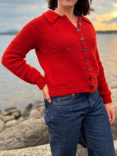 Load image into Gallery viewer, A person wearing a bright red textured knit cardigan with a collar and dark buttons, paired with blue jeans, standing outdoors near water and rocks at sunset.