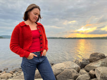 Load image into Gallery viewer, A woman wearing a bright red knit cardigan over a red top and blue jeans stands on a rocky lakeshore at sunset, with water and cloudy sky in the background.