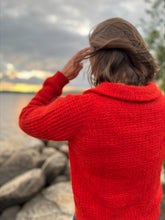 Load image into Gallery viewer, A person viewed from behind wearing a bright red chunky knit sweater, standing on rocks by a body of water under a cloudy sky.