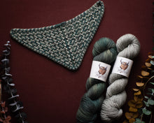 Load image into Gallery viewer, Knitted green and gray triangle scarf with two skeins of yarn on a dark red background